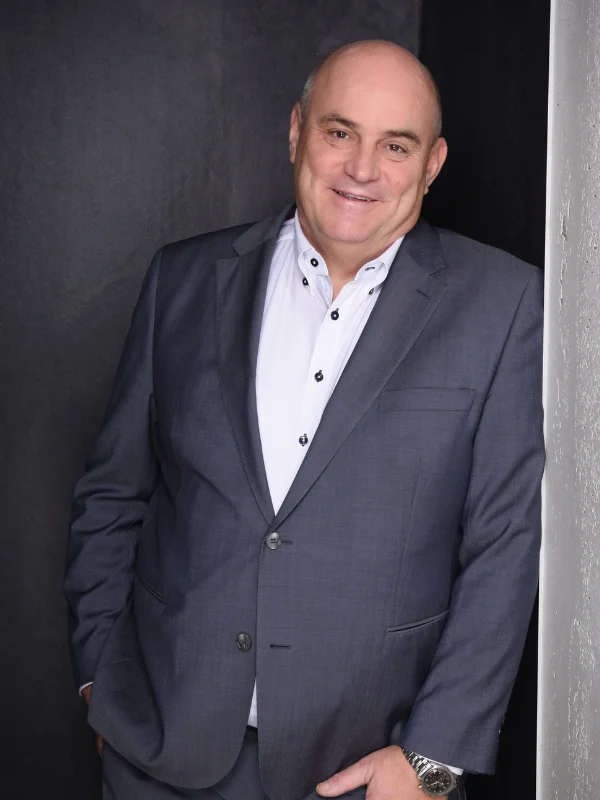 Porträt eines lächelnden Mannes im grauen Anzug vor dunklem Hintergrund, Businessfoto für Lessig Consulting.